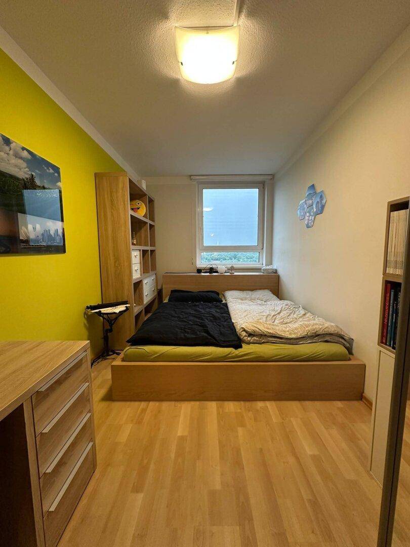 Schlafzimmer mit Holzbett, Regal und einem Fenster, das den Raum mit Tageslicht versorgt.