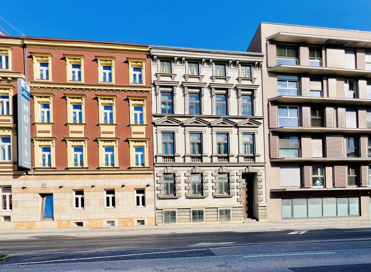 Reihe von Gebäuden unterschiedlicher Architekturstile entlang einer Straße unter blauem Himmel.