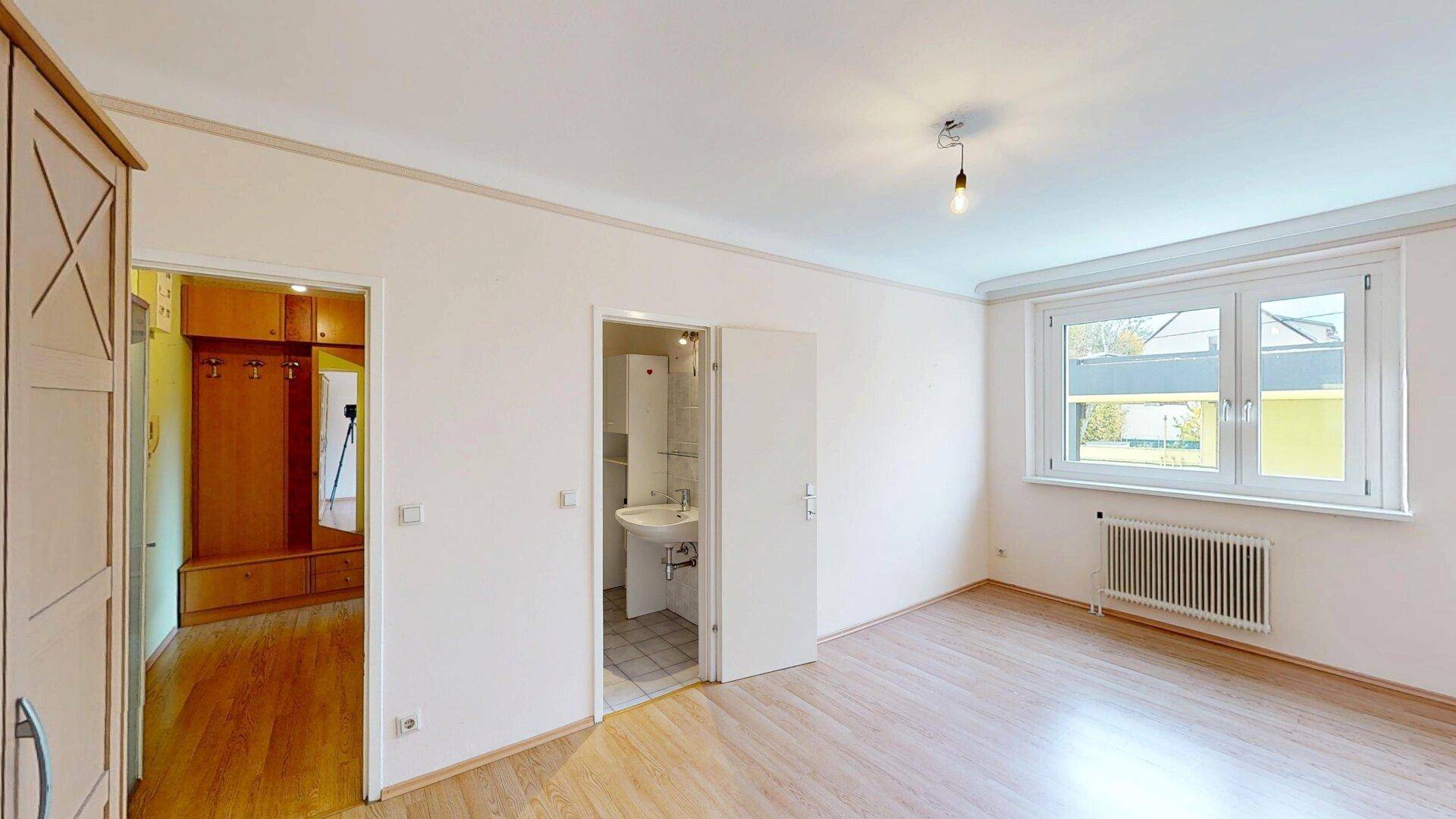 Helles Zimmer mit Parkettboden, weiß gestrichenen Wänden und Zugang zu einem Badezimmer.