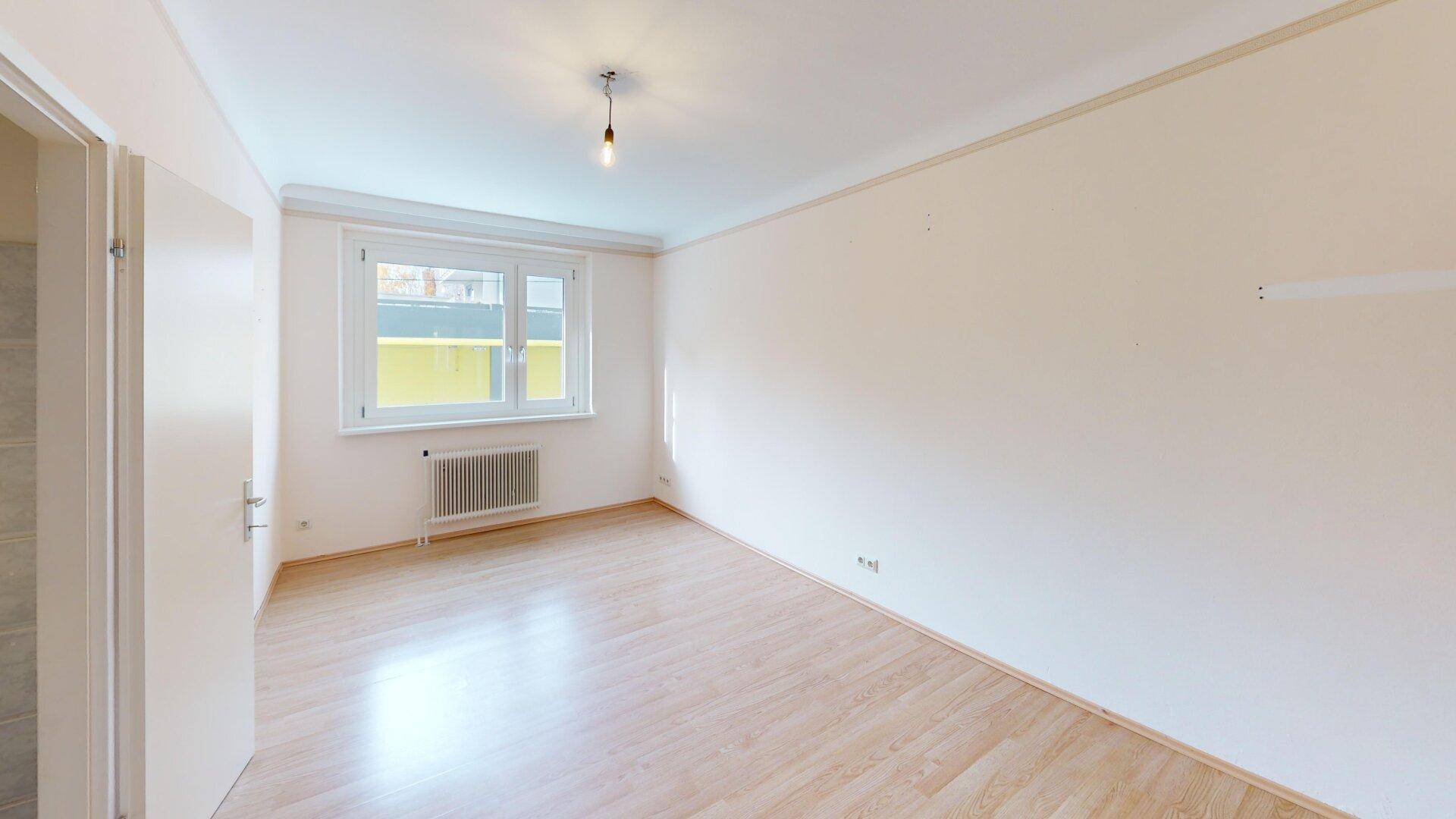 Leeres Zimmer mit Parkettboden, weißen Wänden und einem Fenster mit Blick nach draußen.