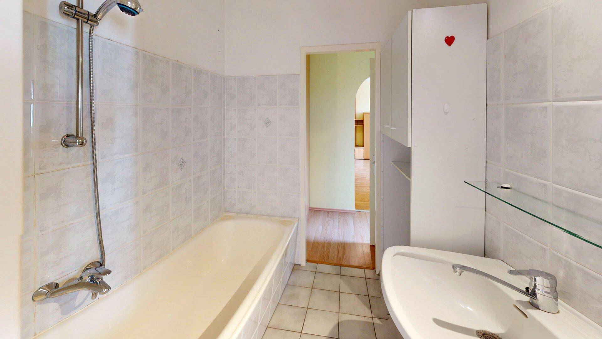 Badezimmer mit Badewanne, Waschbecken und weißen Fliesen, mit Zugang zu einem anderen Raum.