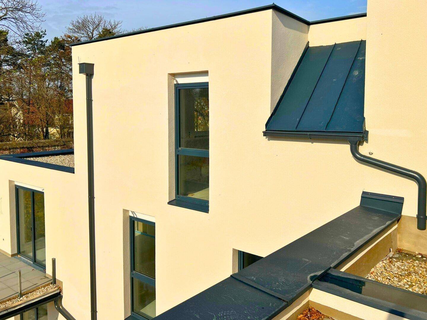 Fassade eines hellbeigen Gebäudes mit dunklen Fensterrahmen und einem dunklen Dachabschnitt.