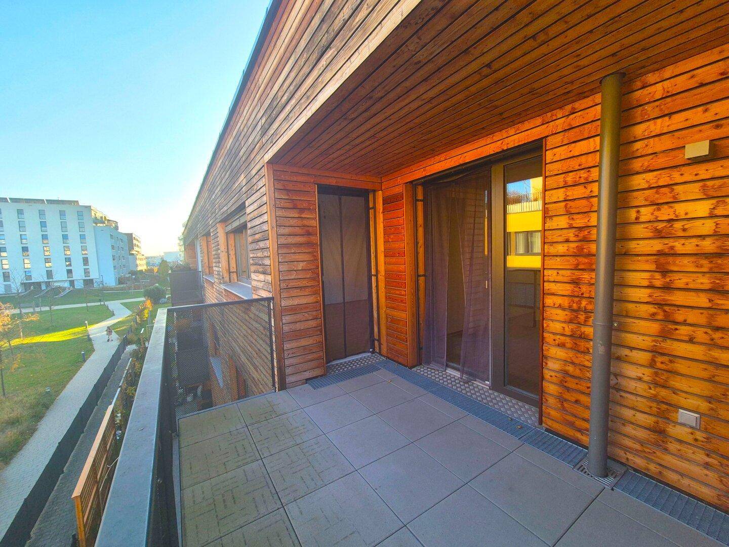 Sonniger Balkon mit Holzfassade, Glasgeländer und Blick ins Grüne.