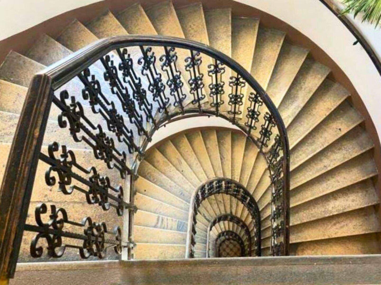 Blick von oben auf die kunstvolle Wendeltreppe mit schmiedeeisernem Geländer und Steinstufen in einem historischen Gebäude.