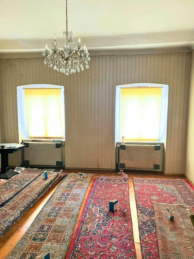 Geräumiger Raum mit Holzdielenboden, zwei Fenstern und einem Kronleuchter, mit Teppichen ausgelegt.