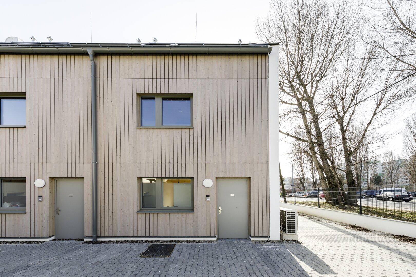 Seitenansicht der Reihenhäuser mit Holzfassade und gepflastertem Zugangsbereich.