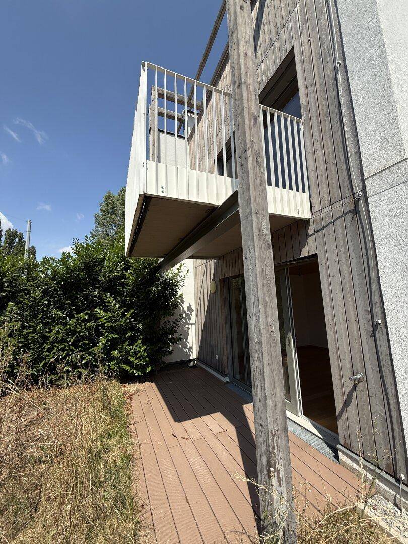 Gartenbereich mit Holzterrasse und Balkon darüber, umgeben von Hecken.