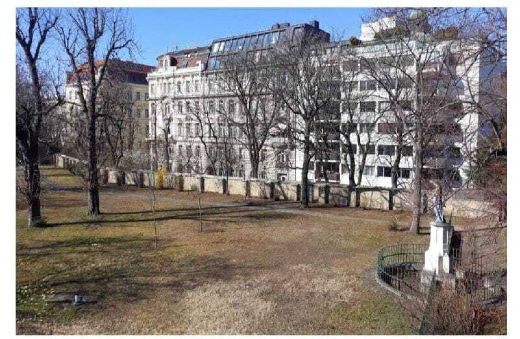 Grüne Parkanlage mit hohen Bäumen und historischen Gebäuden im Hintergrund.