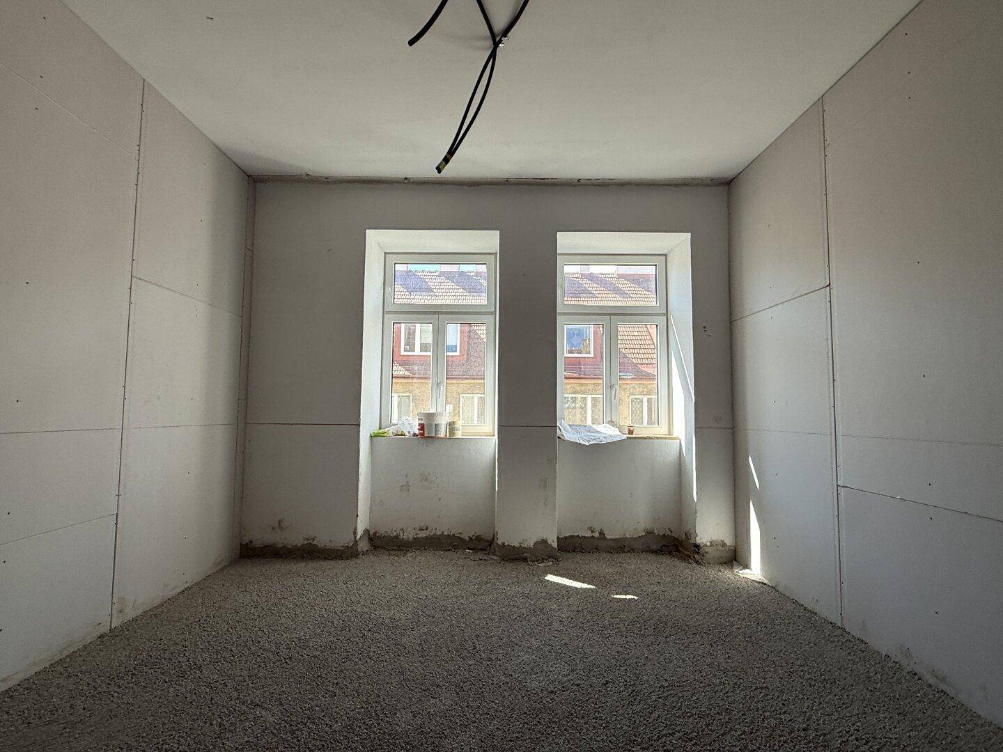 Unfertiger Raum mit zwei Fenstern, die Tageslicht hereinlassen, und vorbereitetem Estrichboden.