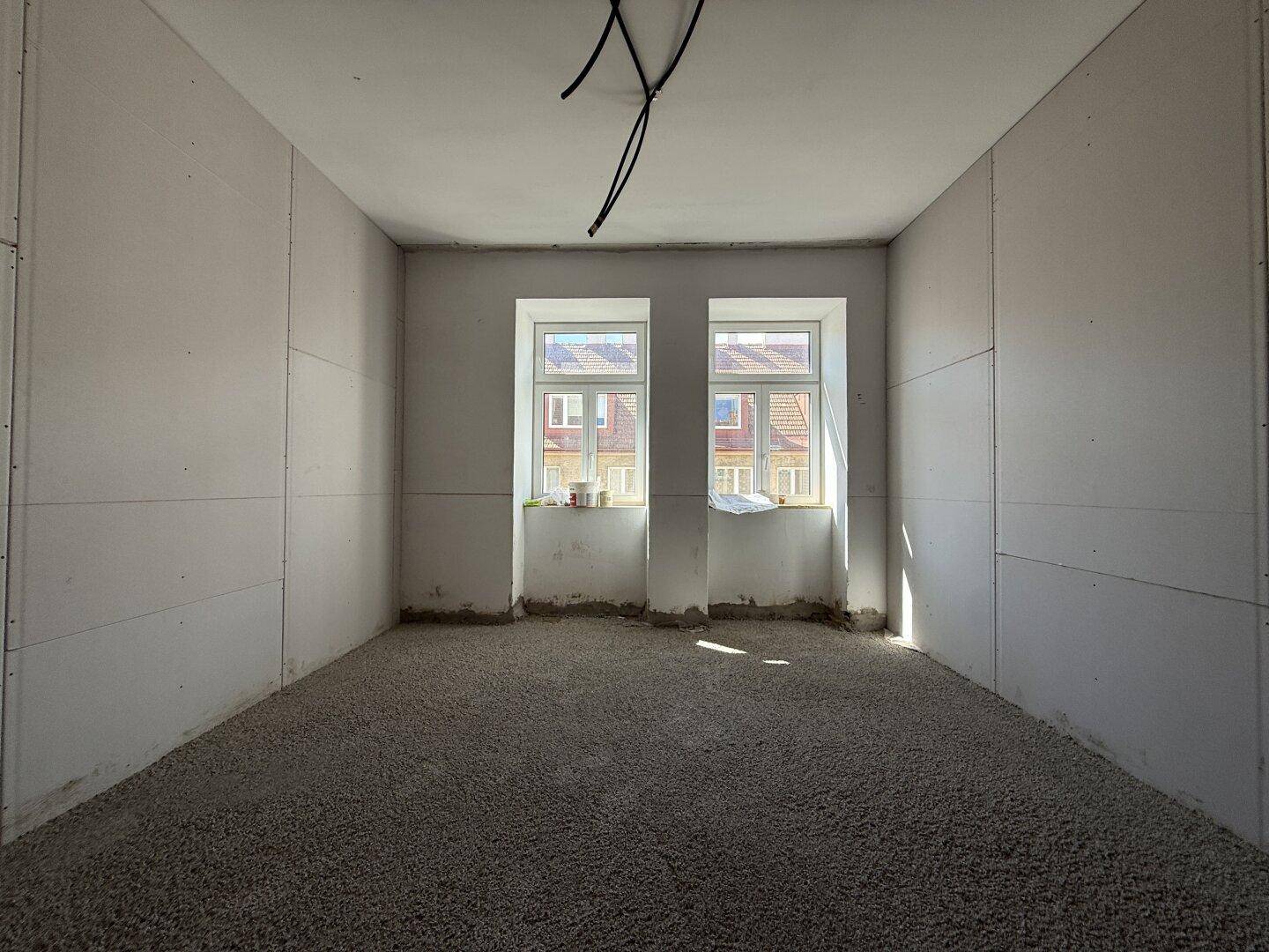 Heller Raum im Rohbau mit zwei Fenstern und vorbereitetem Boden für den weiteren Ausbau.