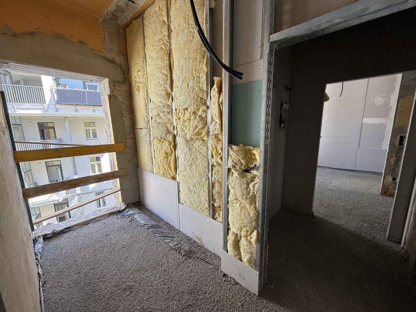 Baustelle mit Dämmmaterial an den Wänden und Blick auf benachbarte Gebäude durch ein Fenster.