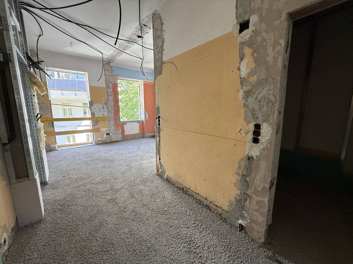 Großer Raum im Rohbau mit freigelegten Wänden und Blick auf die Umgebung durch Fenster.