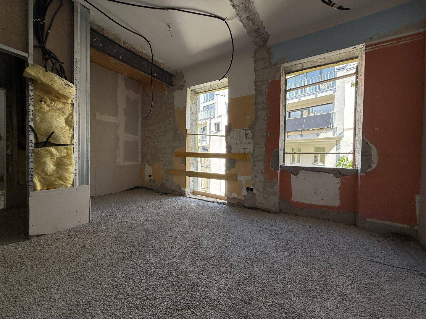 Raum im Rohbau mit freigelegten Wänden, vorbereitetem Boden und Blick nach draußen.