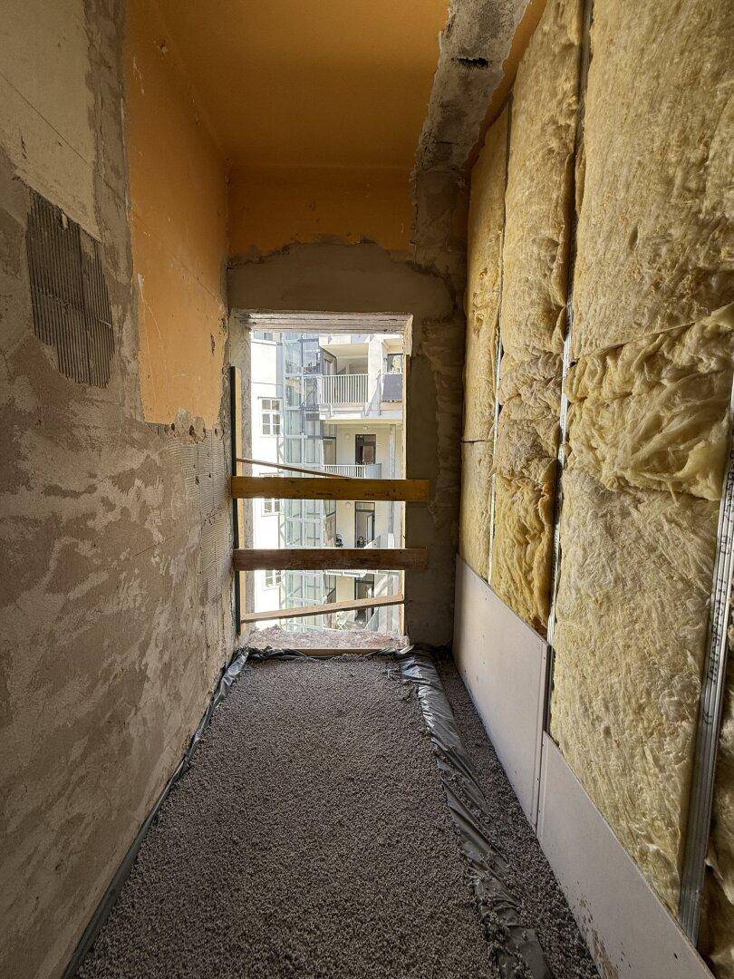 Schmaler Bereich im Rohbau mit Dämmung und Blick auf einen Innenhof durch das Fenster.