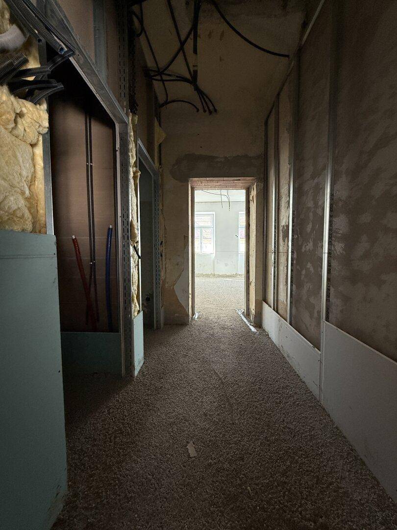 Langer Flur im Rohbau mit sichtbaren Installationen und Blick in einen weiteren Raum.