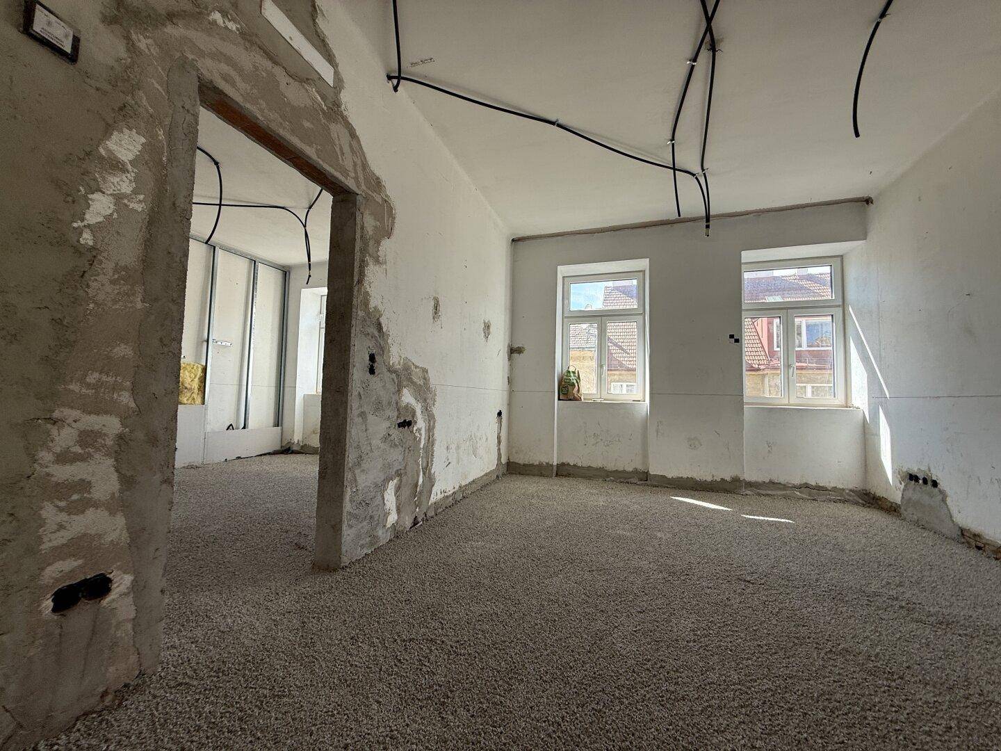 Großer Raum im Rohbau mit freigelegten Wänden, sichtbaren Leitungen und zwei Fenstern.