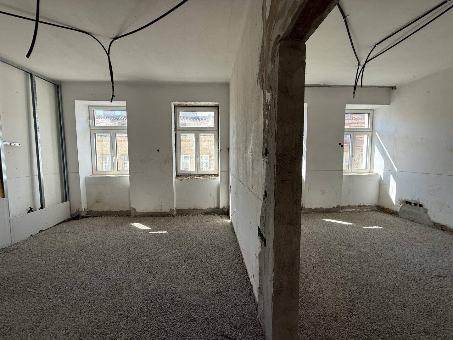Großer Raum im Rohbau, durch eine Wand geteilt, mit zwei Fenstern und vorbereitetem Boden.