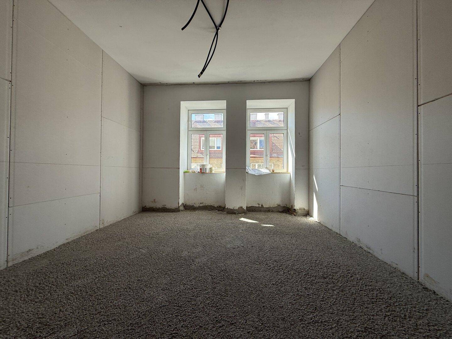 Unfertiger Raum mit zwei Fenstern, die Tageslicht hereinlassen, und vorbereitetem Estrichboden.