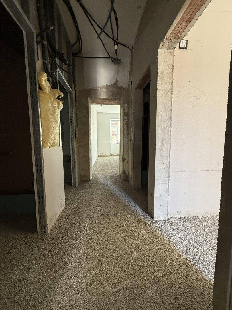 Langer Flur im Rohbau mit sichtbaren Installationen und Blick in einen weiteren Raum.