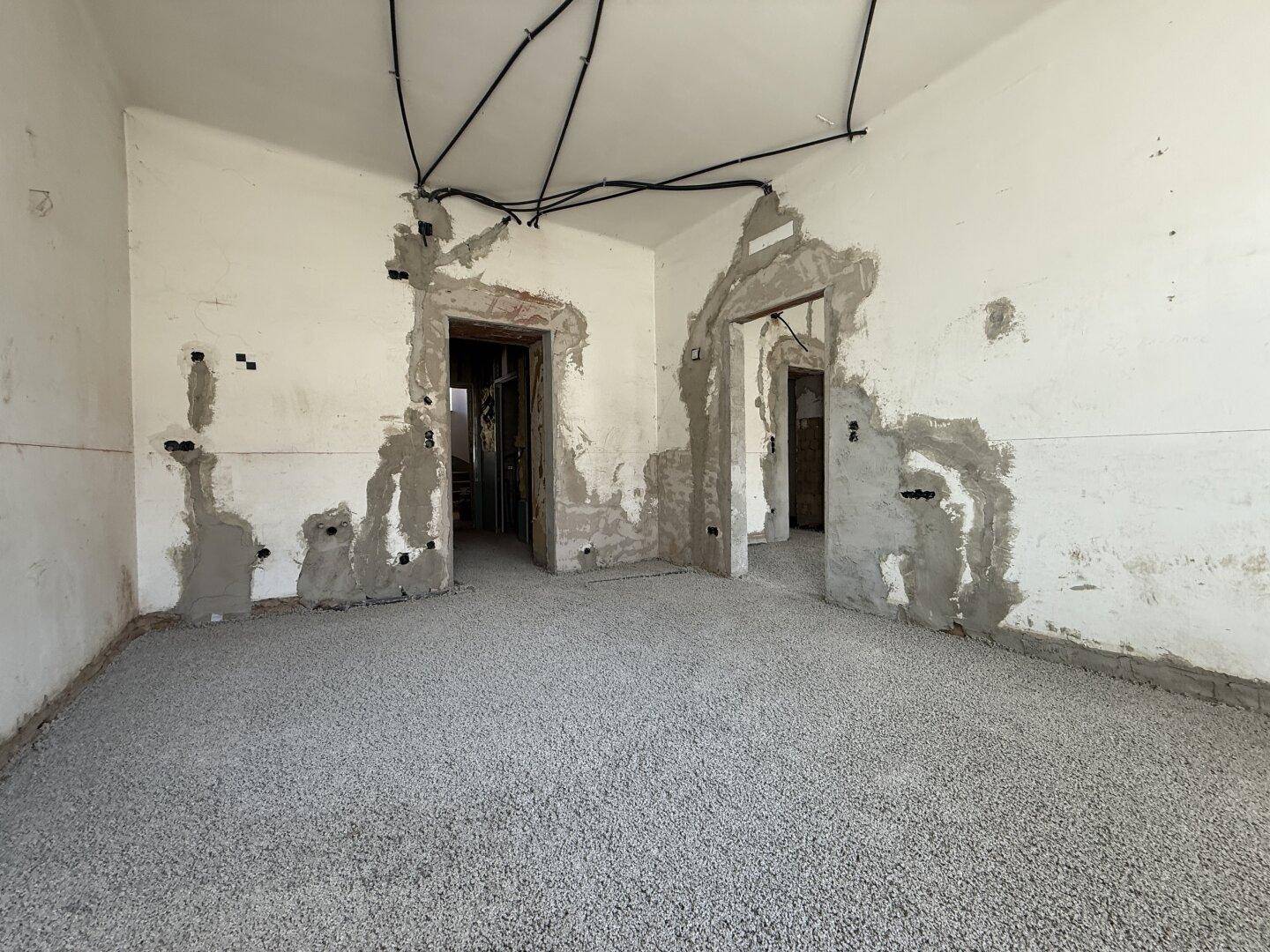 Raum im Rohbau mit freigelegten Wänden, sichtbaren Leitungen und vorbereitetem Boden.