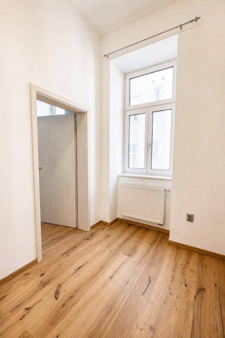 Leerer Raum mit hellem Holzfußboden und einem Fenster, das den Blick nach draußen freigibt.