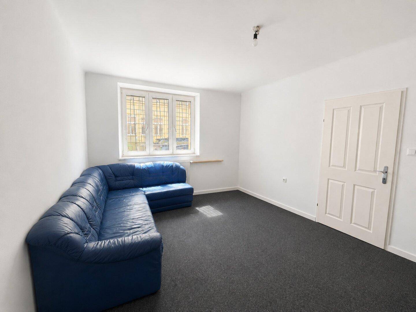 Wohnzimmer mit einem blauen Ecksofa und dunklem Teppichboden, hell durch ein Fenster.