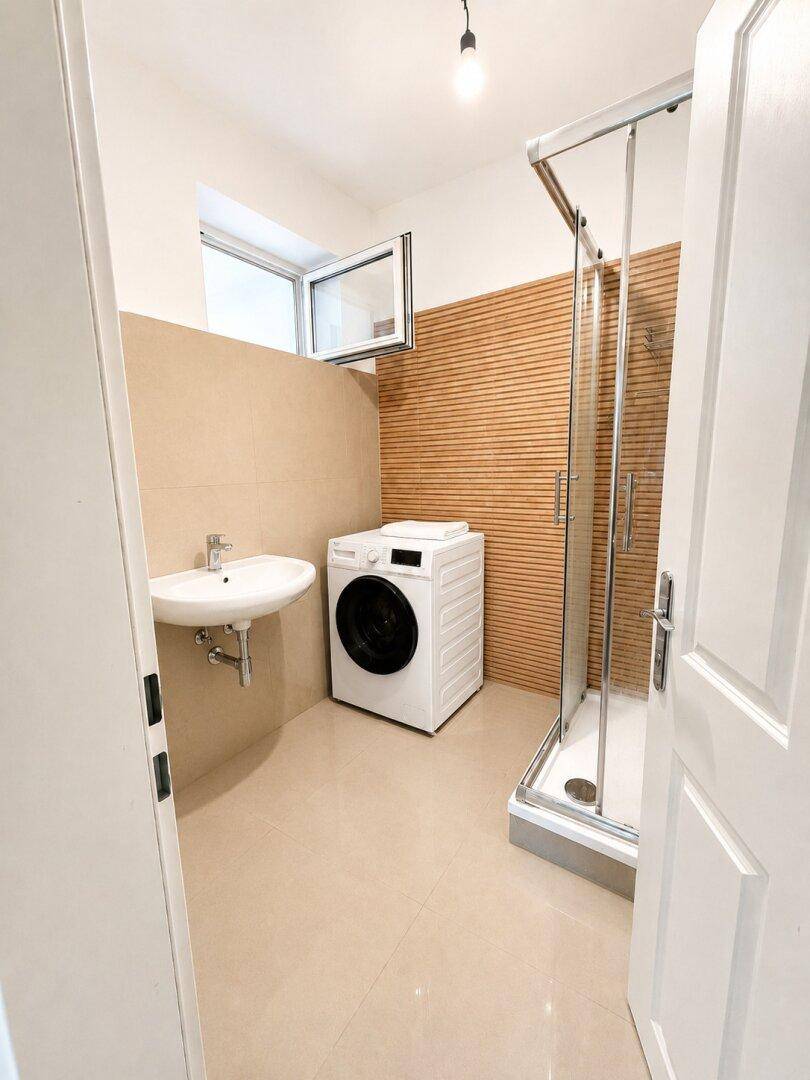 Badezimmer mit Duschkabine, Waschbecken und Waschmaschine, hell durch ein Fenster.