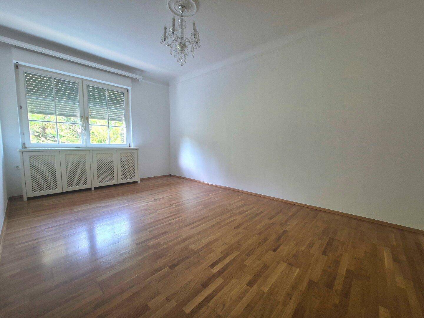 Heller Raum mit Parkettboden, Fenster mit Rollläden und einem eleganten Kronleuchter an der Decke.