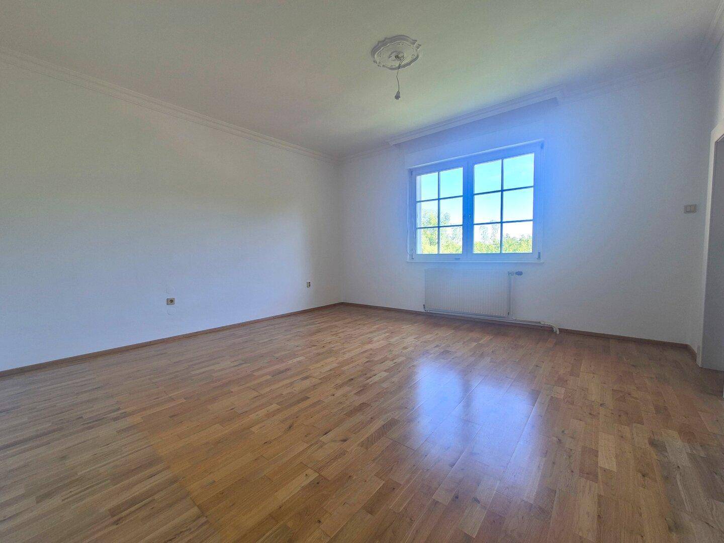 Geräumiger Raum mit hellem Parkettboden, einem großen Fenster und Stuckelementen an der Decke.