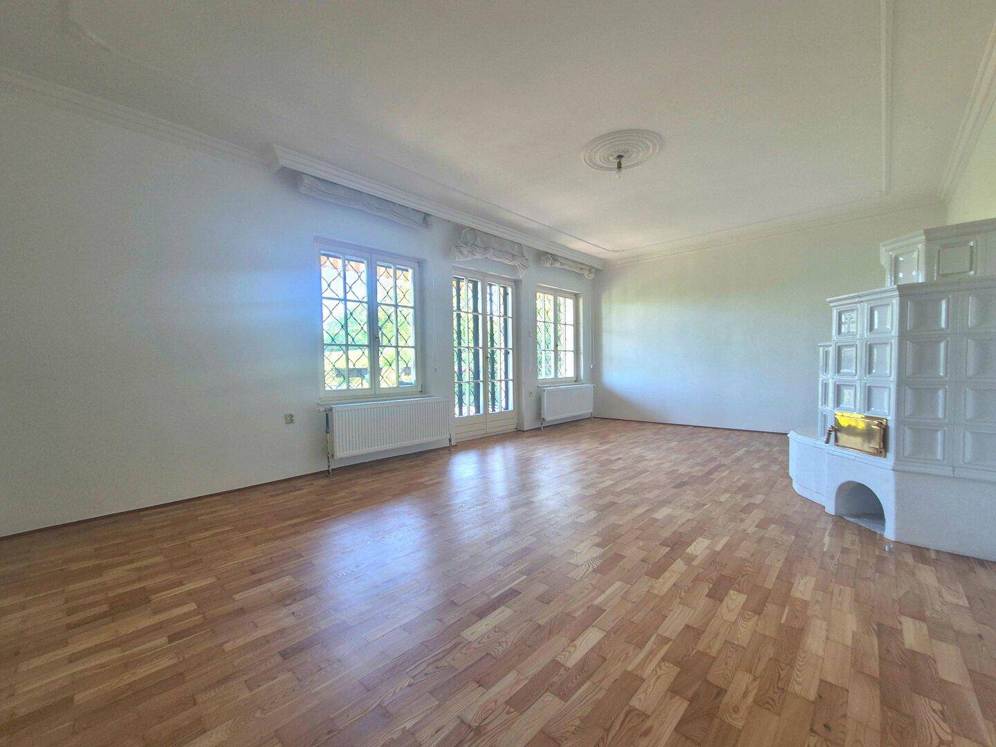 Helles Wohnzimmer mit Parkettboden, großen Fenstern und einem traditionellen Kachelofen.