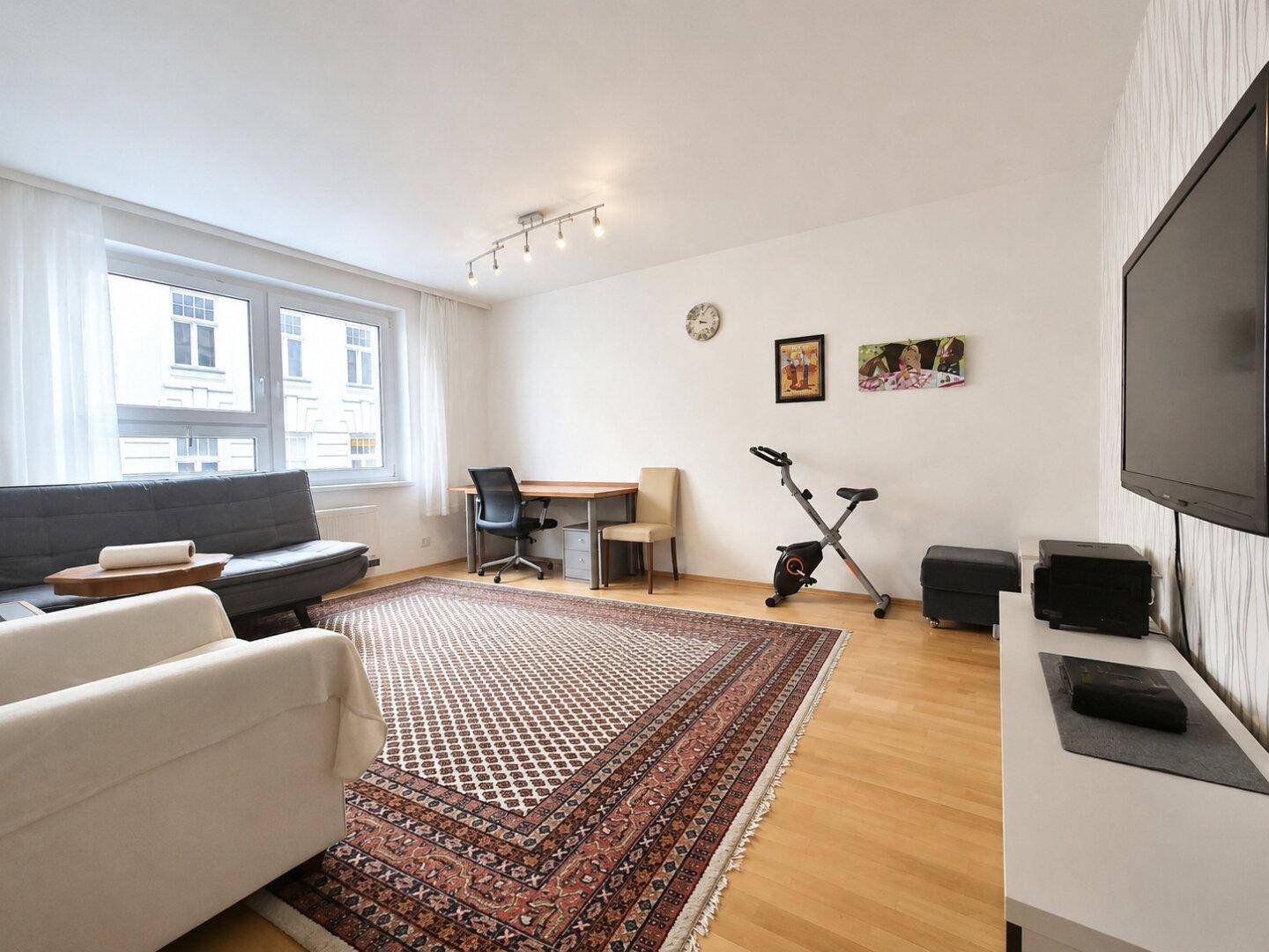 Helles Wohnzimmer mit Laminatboden, großem Teppich, Schreibtisch und einem Fitnessgerät.