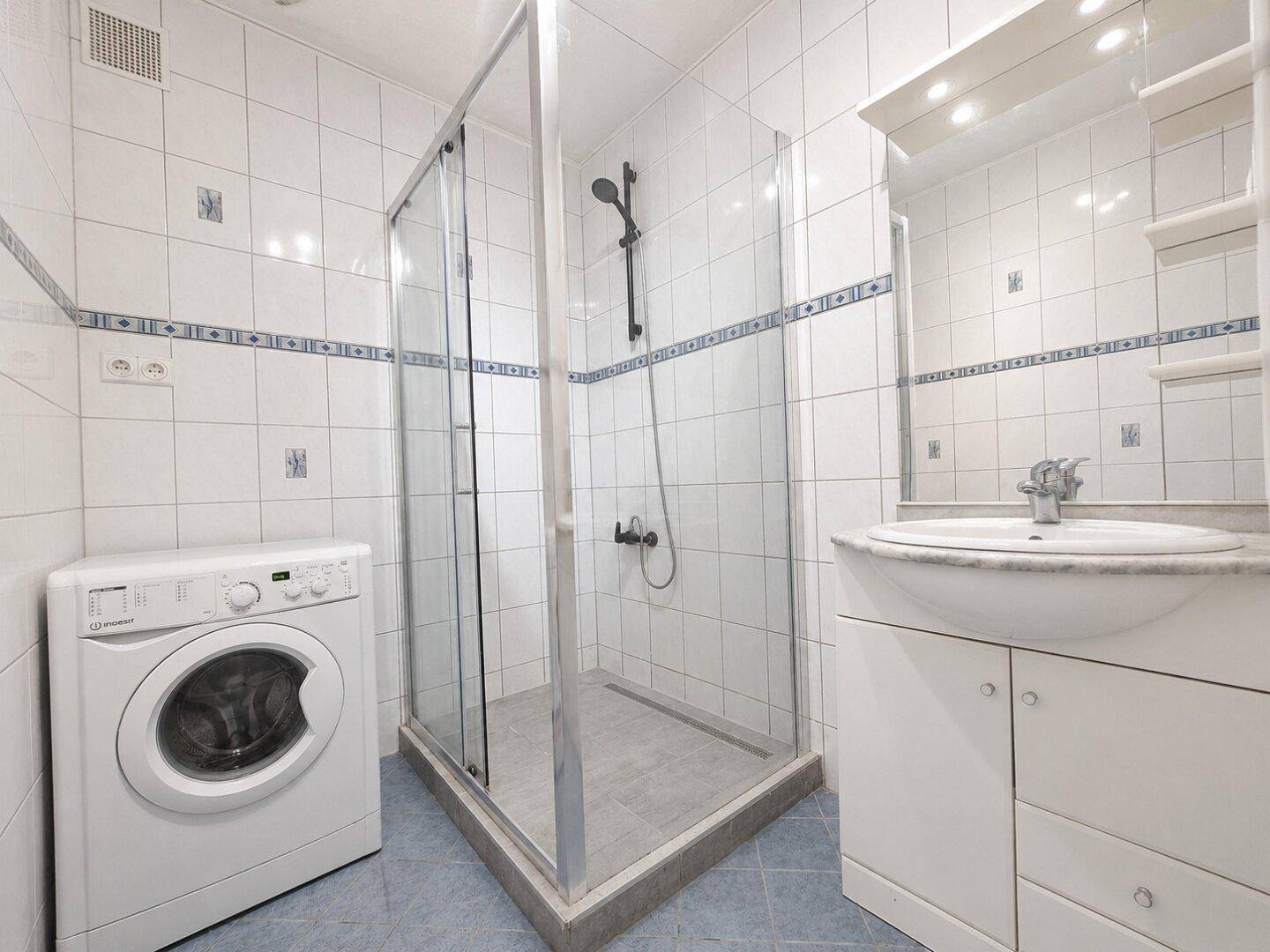 Badezimmer mit Duschkabine, Waschbecken und Waschmaschine, ausgestattet mit weißen Fliesen und blauen Akzenten.