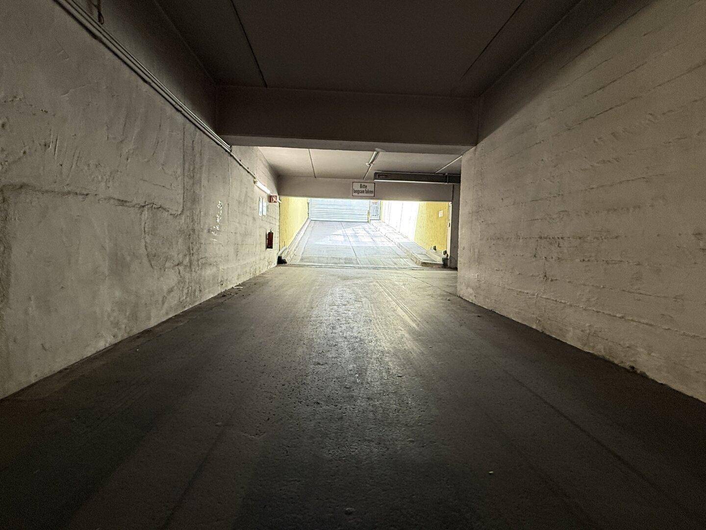 Blick aus einem Tiefgaragentunnel mit Betonwänden, der zum hellen Eingangsbereich führt.
