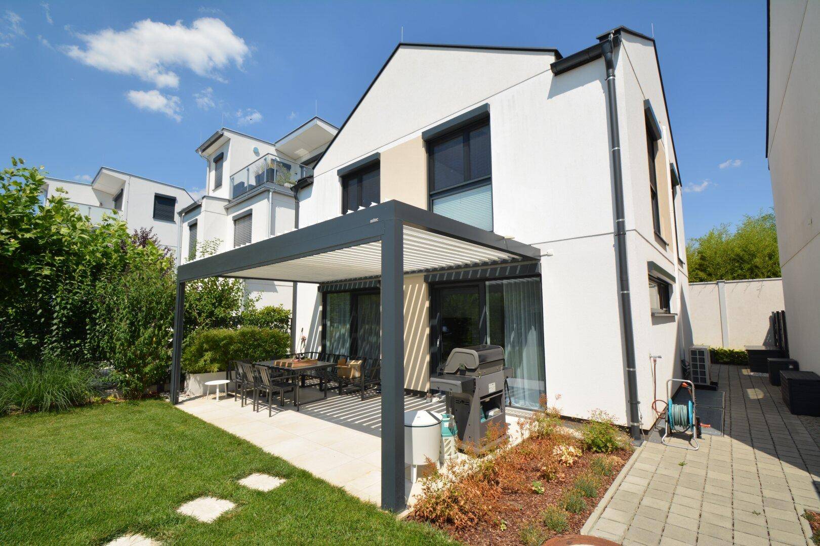Rückseite des modernen Hauses mit Terrasse, Pergola und angrenzendem grünen Gartenbereich.