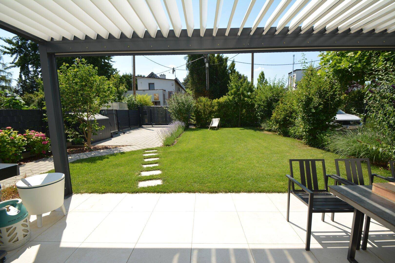 Blick von der überdachten Terrasse in den sonnigen, grünen Garten mit Liegestuhl und Pflanzen.