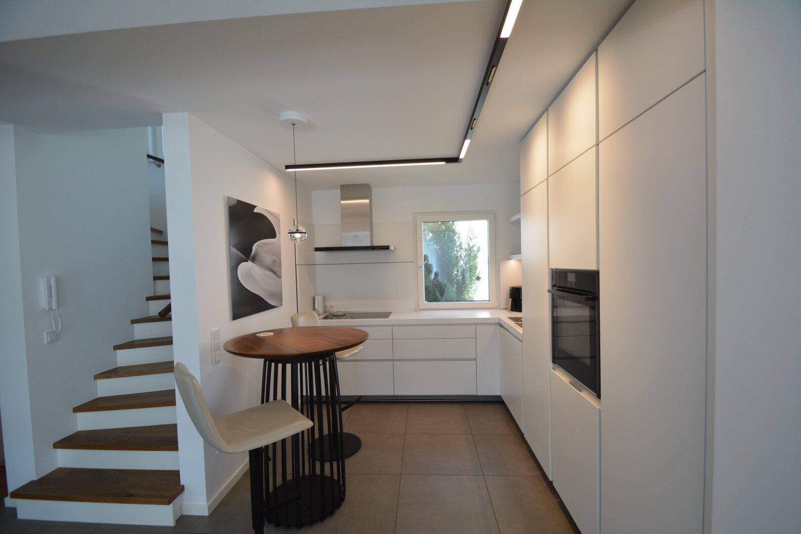 Moderne weiße Küche mit Einbaugeräten, Kochinsel und angrenzender Holztreppe.