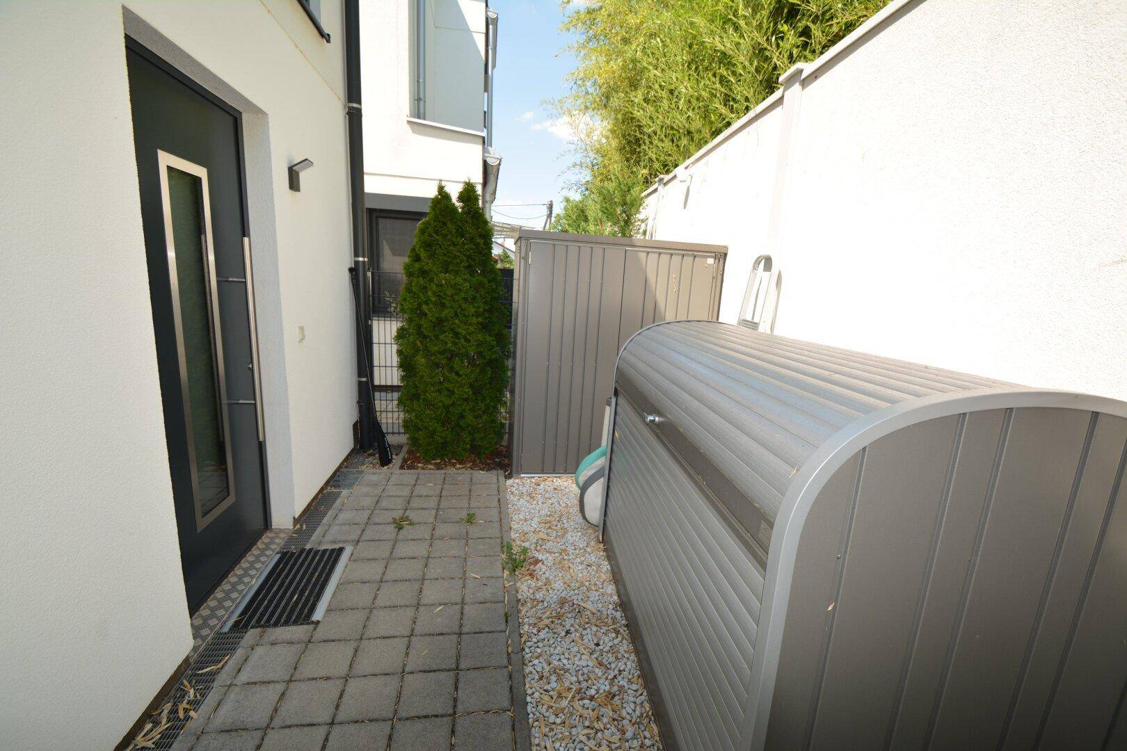 Seitlicher Zugang zum Haus mit Eingangstür, gepflastertem Weg und diskreten Mülltonnenboxen.