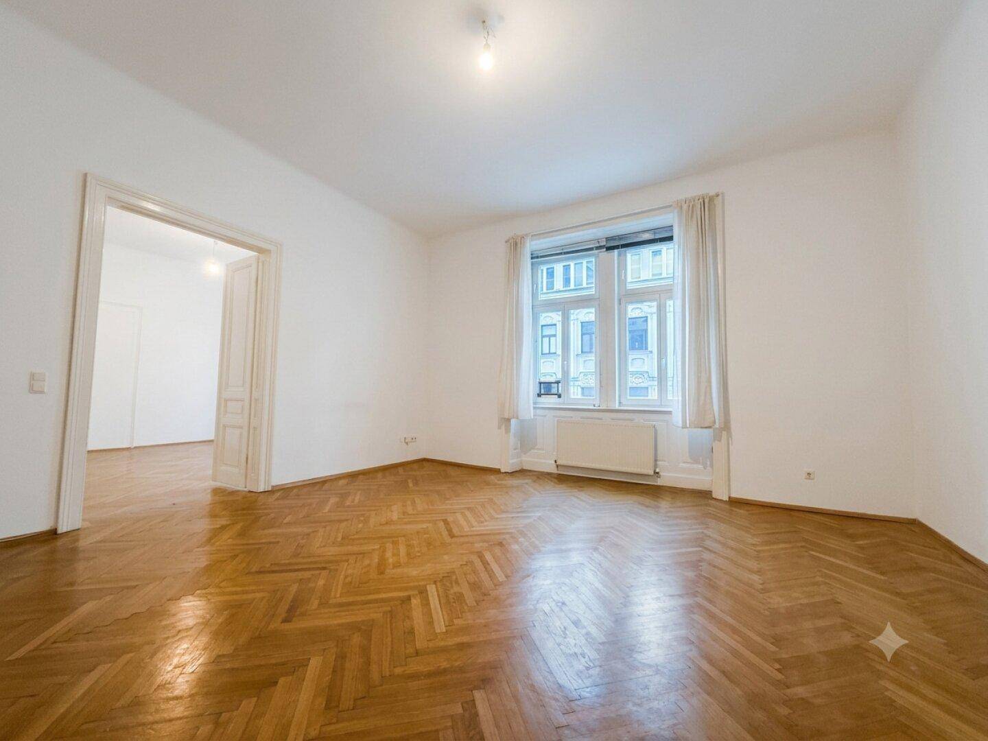 Geräumiges Zimmer mit klassischem Fischgrätparkett und großen Fenstern, ideal als Wohn- oder Schlafzimmer.
