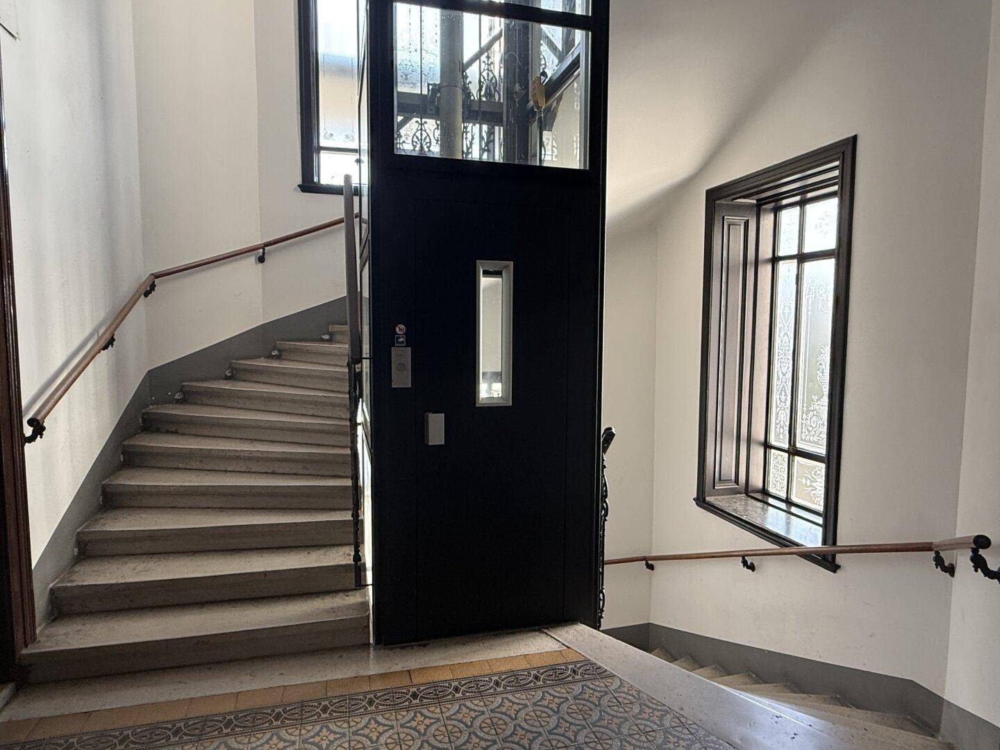 Historisches Treppenhaus mit elegantem Aufzug und originalen Fliesenböden in einem Altbau.