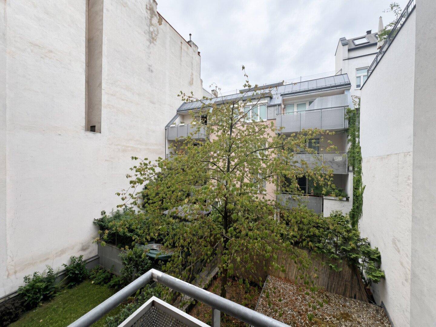 Blick vom Balkon in einen grünen Innenhof mit Bäumen und umliegenden Gebäuden.