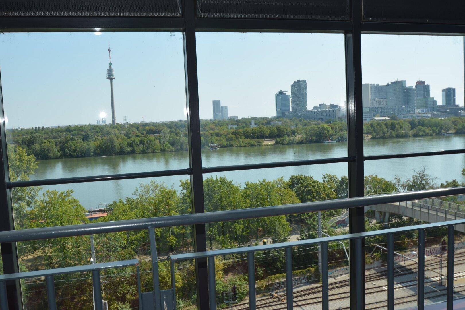 Panoramablick auf die Donau, die Wiener Skyline und den Donauturm durch große Fenster.