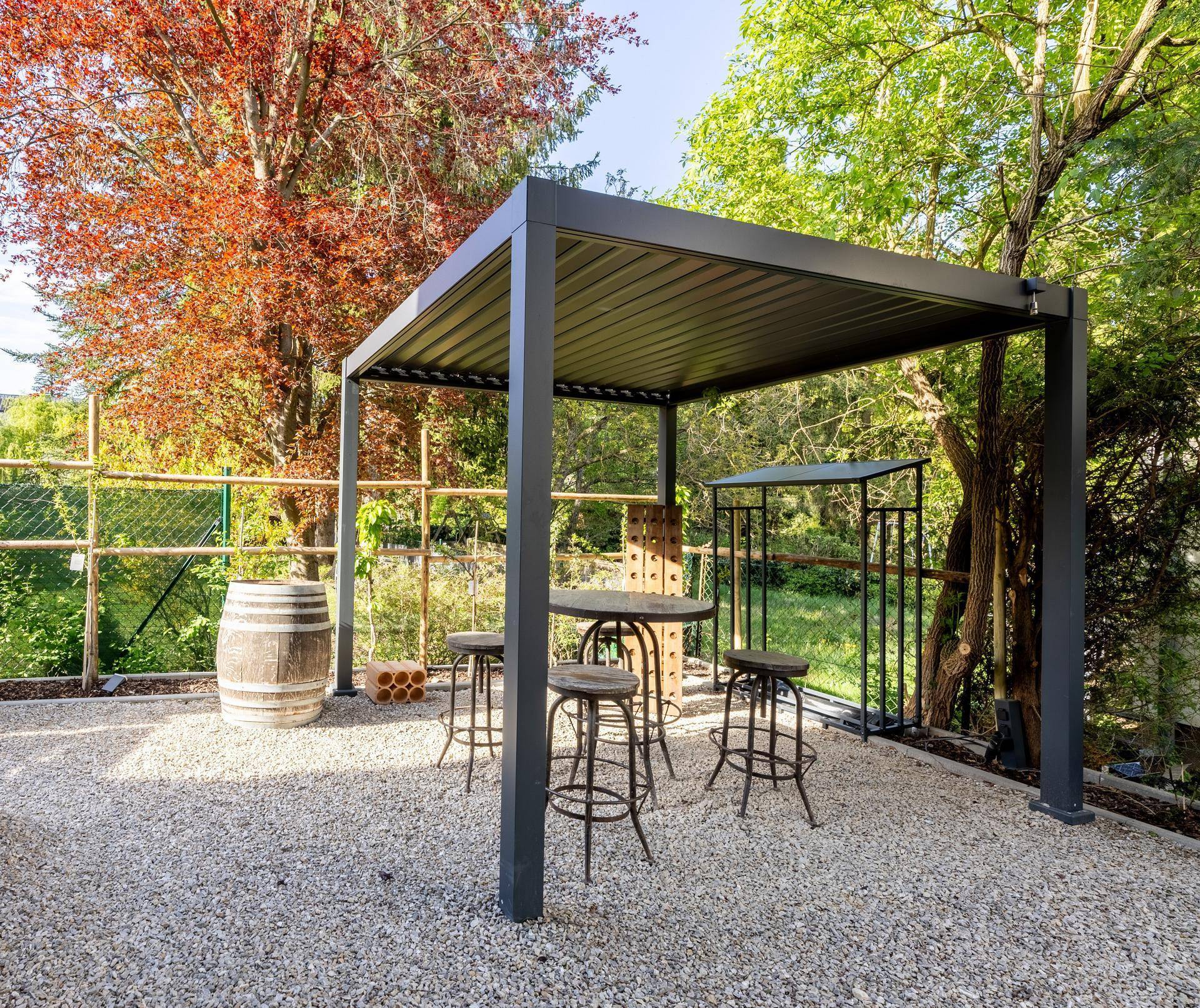 Gemütlicher Sitzbereich unter einer Pergola mit Bar und Fass auf Kiesfläche im Garten.