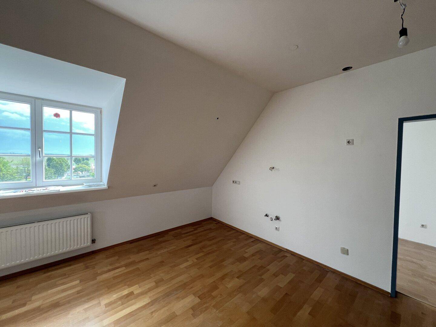 Helles Zimmer im Dachgeschoss mit Holzfußboden, Dachschräge und großem Fenster.