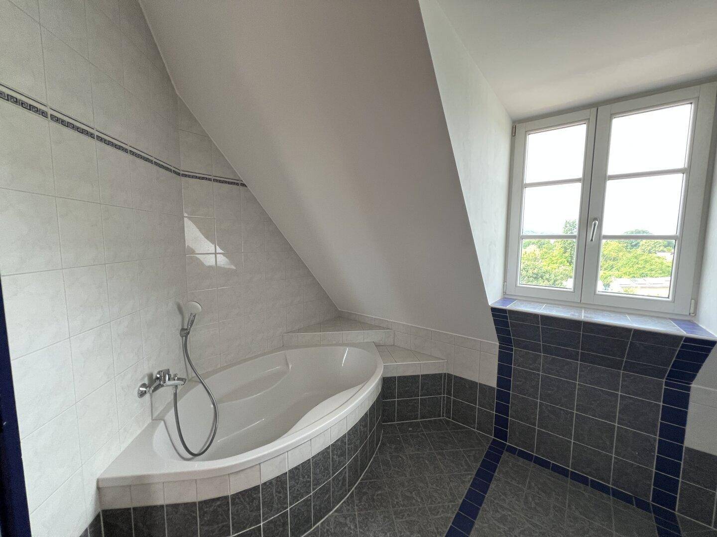 Eckbadewanne im hellen Badezimmer mit Fliesen, Dachschräge und Fenster mit Ausblick.