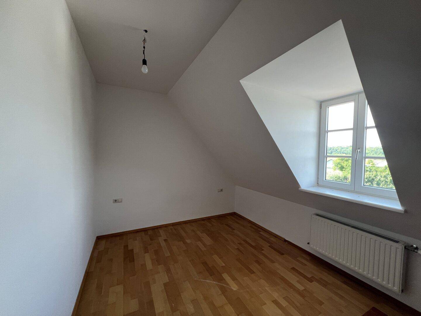 Helles Dachgeschosszimmer mit Holzfußboden, Dachschräge und Fenster.