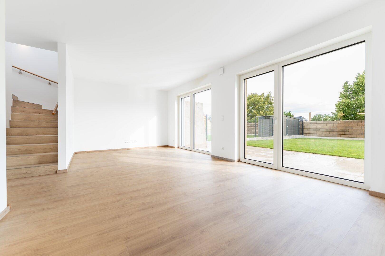 Helles, leeres Wohnzimmer mit Laminatboden, großen Fenstern und Zugang zum Garten.