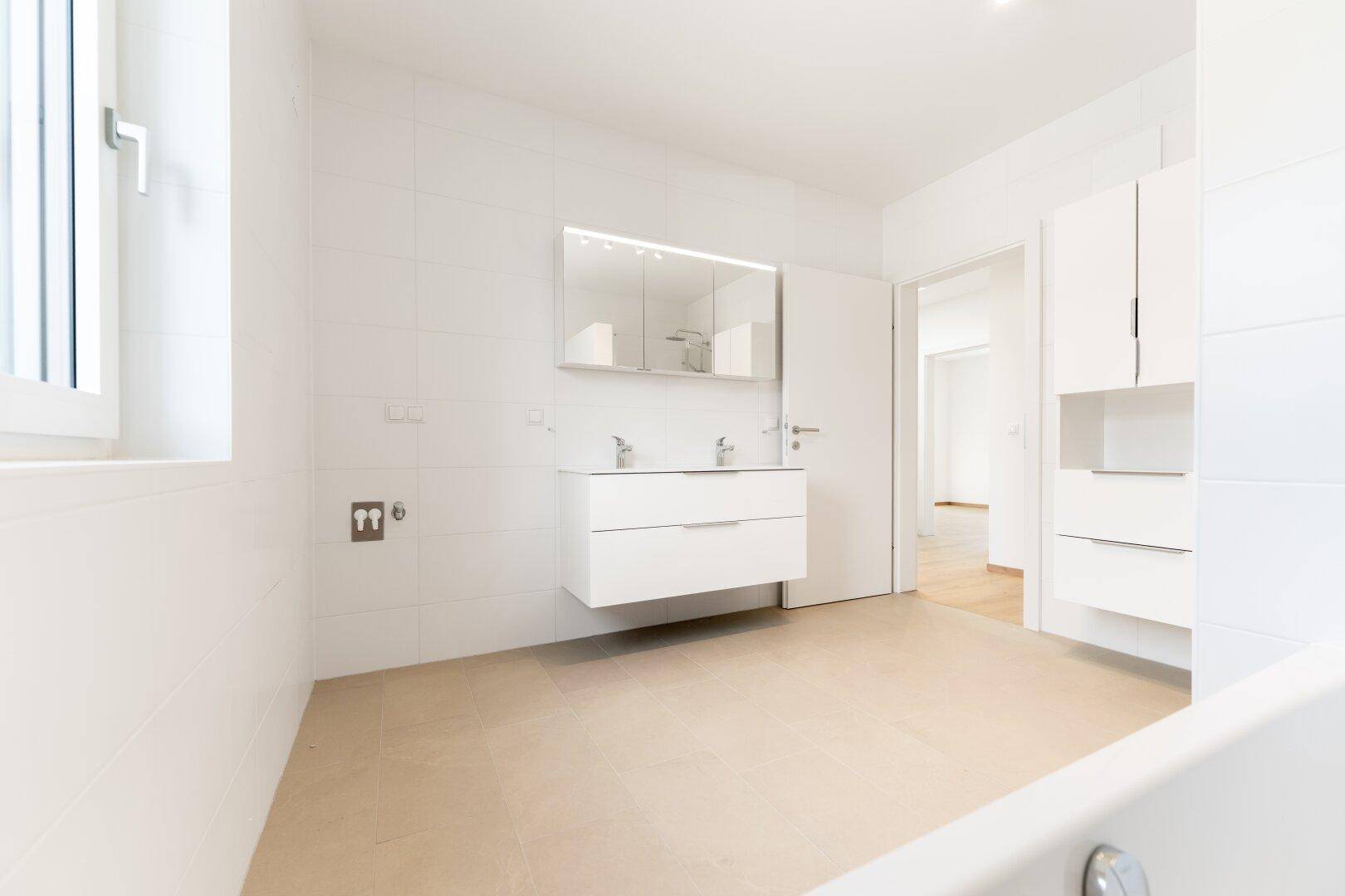 Geräumiges Badezimmer mit weißen Fliesen, Doppelwaschbecken, Spiegelschrank und Fenster.