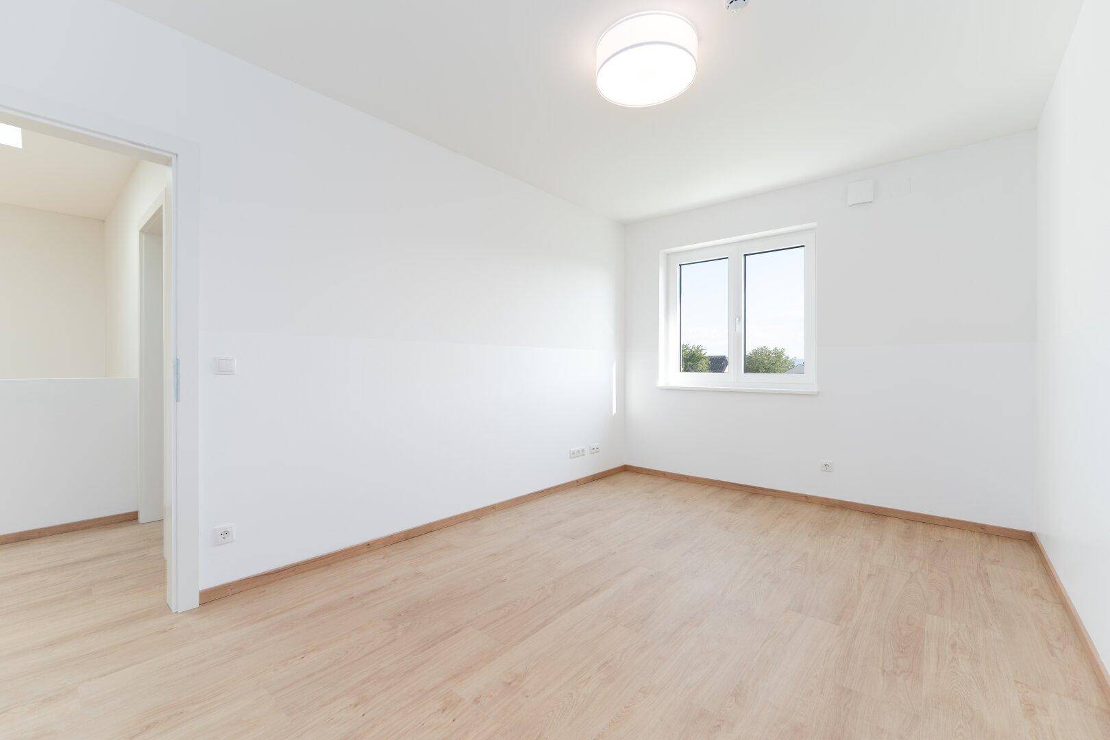 Leeres Schlafzimmer mit hellem Laminatboden, weißen Wänden und einem Fenster mit Ausblick.