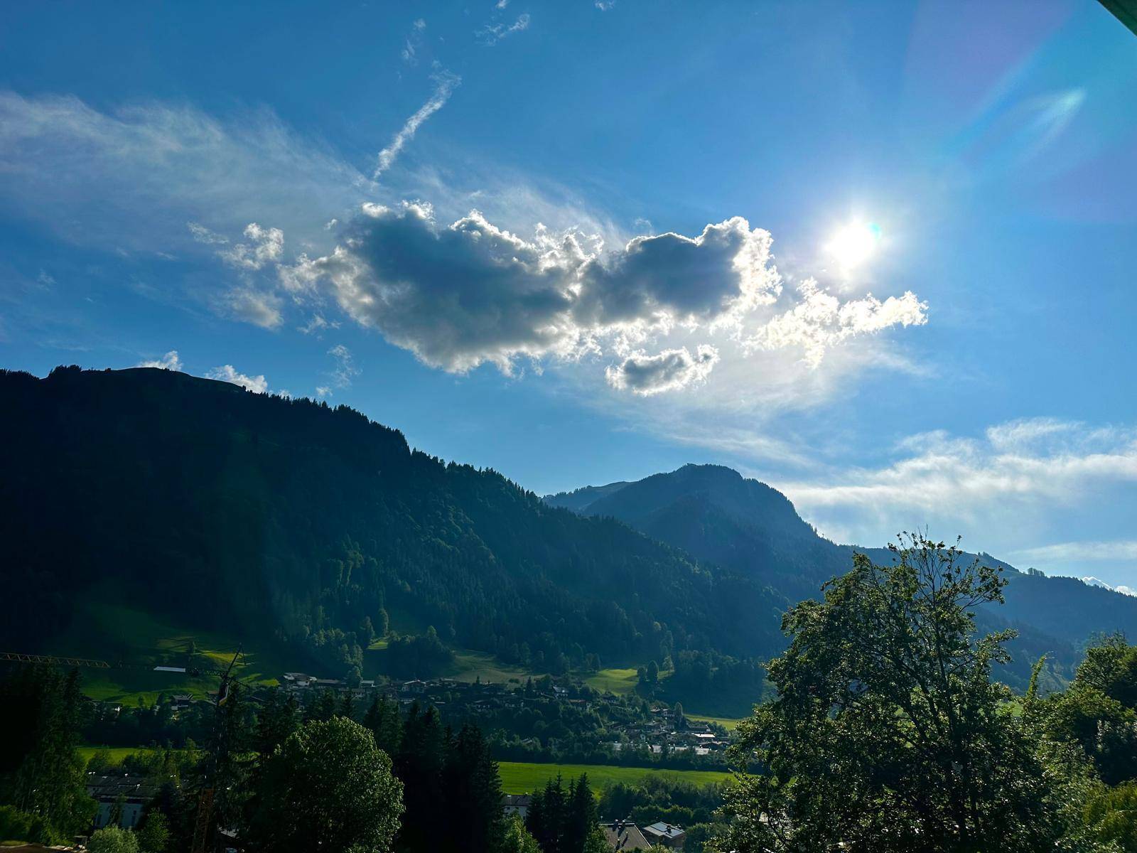 Sonnige Berglandschaft im Sommer mit grünen Hügeln, Bäumen und einem Dorf im Tal.