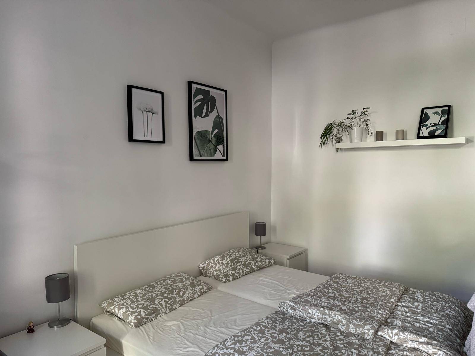 Helles Schlafzimmer mit Doppelbett, Nachttischen und schlichten Wanddekorationen auf Holzboden.